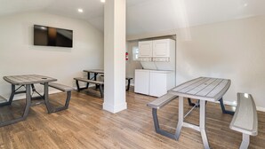 Un interior moderno con un televisor de pantalla plana montado en la pared, una kitchenette con microondas y refrigerador pequeño, y tres mesas tipo picnic con bancos.