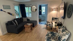 Ein Wohnzimmer mit einem dunkelgrauen Sofa, einem Holztisch und einem blumigen Sessel. An der Wand hängt ein Spiegel und am Fenster hängen blaue Vorhänge.
