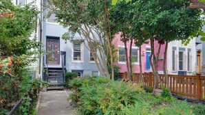 A narrow pathway flanked by lush greenery and a wooden fence, leading to a row of colorful townhouses with visible windows and doors. - Central DC Location! Urban Oasis in Logan Circle (Washington)