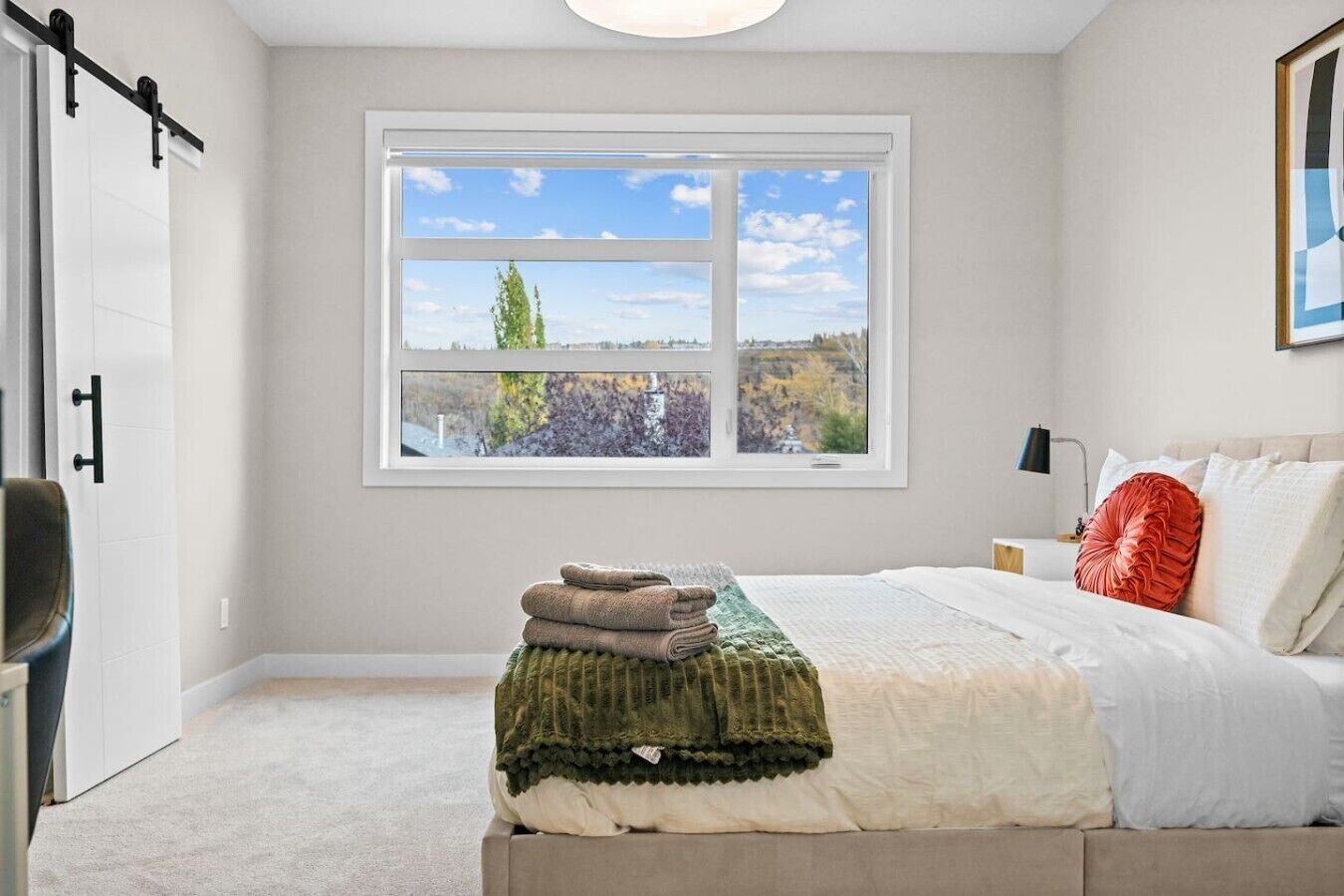 A bedroom with a large window, a bed with pillows, a bedside table with a lamp, and a chair.