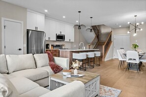 Una cocina moderna con armarios blancos, un refrigerador de acero inoxidable y un microondas. Hay un comedor con sillas y una mesa blancos, y una escalera que lleva a un nivel superior.