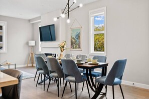 Uma zona de refeições moderna com uma mesa posta para quatro pessoas, rodeada por seis cadeiras, uma televisão de ecrã plano na parede e duas janelas com quadros.