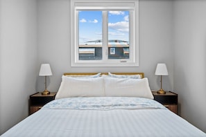 Quarto com cama, duas luminárias de cabeceira, janela com vista e quadro pendurado na parede.