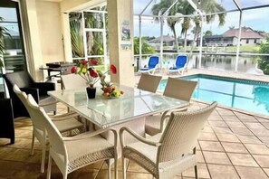 Un tavolo con piano in vetro, sedie in vimini, un vaso di fiori e vista su una piscina e palme.