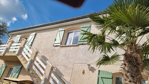 A two-story building with a staircase leading to the upper floor, green shutters on the windows, and a palm tree in the foreground.