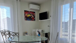 A dining area with a glass table and white metal chairs, a mounted air conditioner, and a television on a shelf.