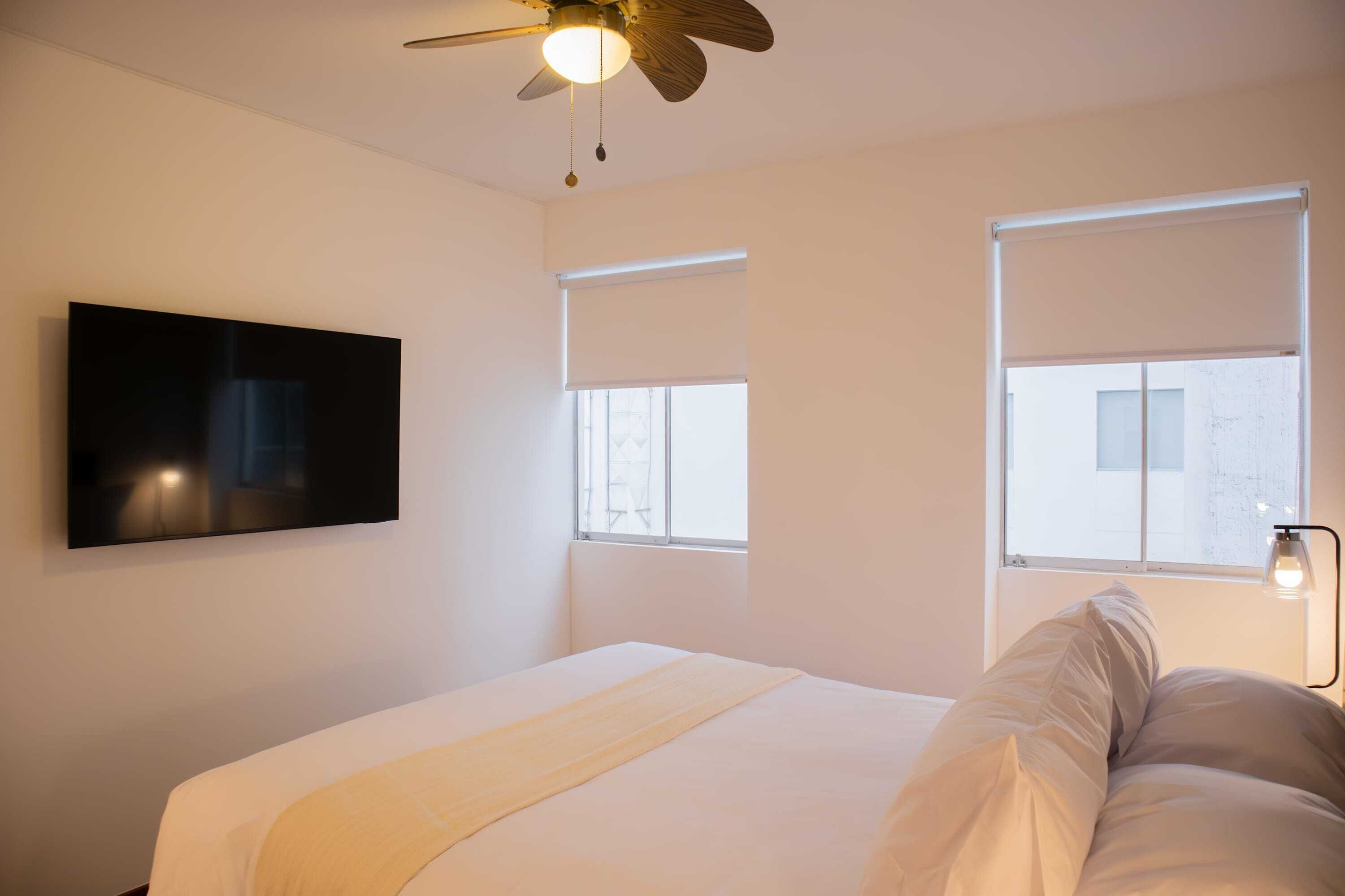 A bedroom with a bed, a ceiling fan, a mounted TV, and two windows with blinds.