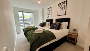 A modern hotel room with a large bed, two side beds, a small bedside table, and a view of the city through a large window. - Stylish 2BR Apt Bracknell | Near Ascot (Bracknell)