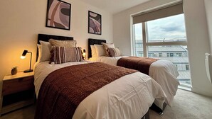A modern bedroom with two beds, a nightstand with a lamp, and a large window offering a view of buildings.