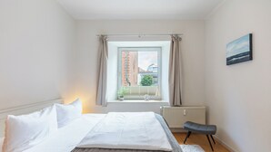 Un dormitorio con cama, ventana con cortinas, radiador, una pequeña banca y un cuadro en la pared.