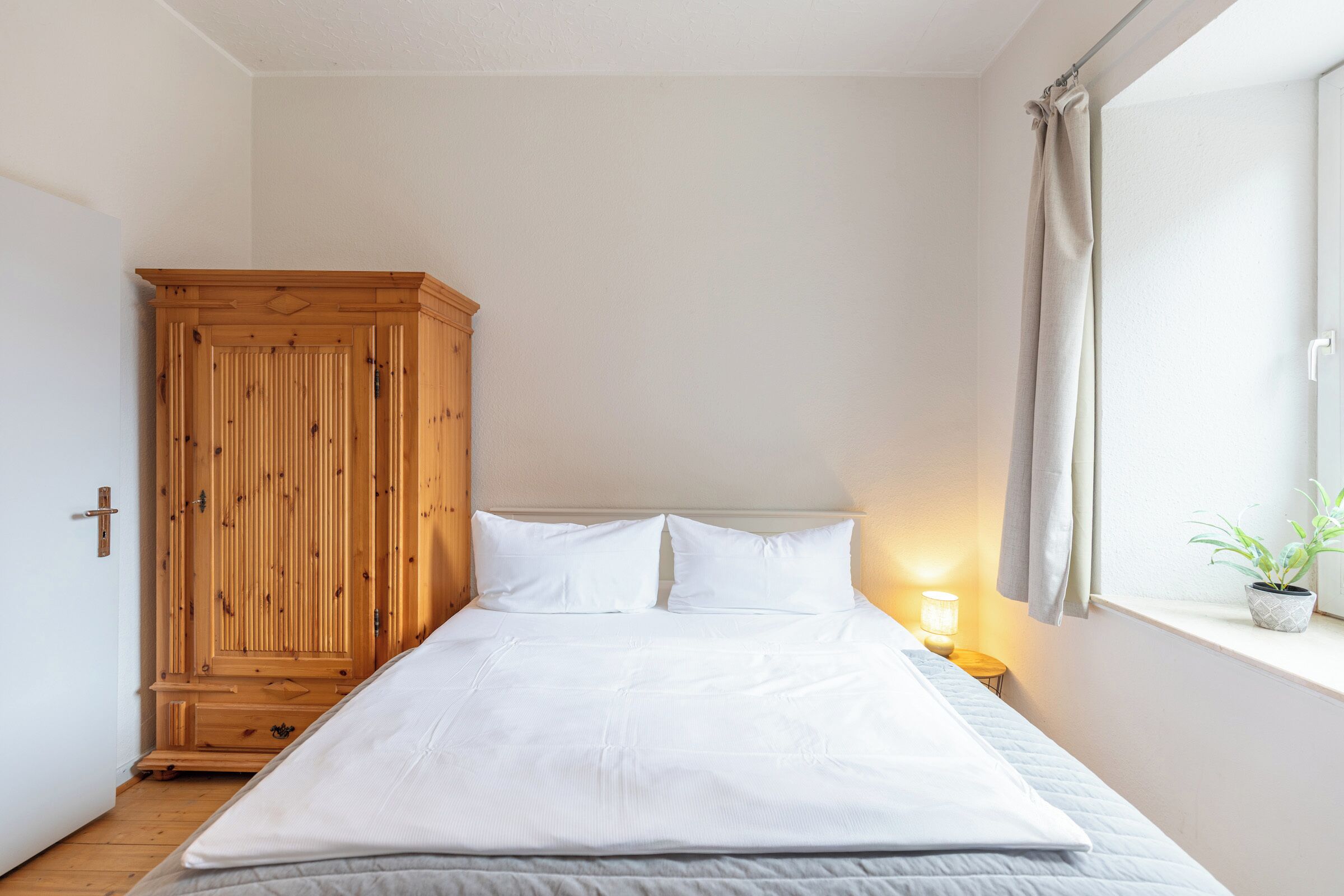 Ein Schlafzimmer mit einem großen Bett, einem Holzschrank, einem Nachttisch mit Lampe und einem Fenster mit Pflanze.