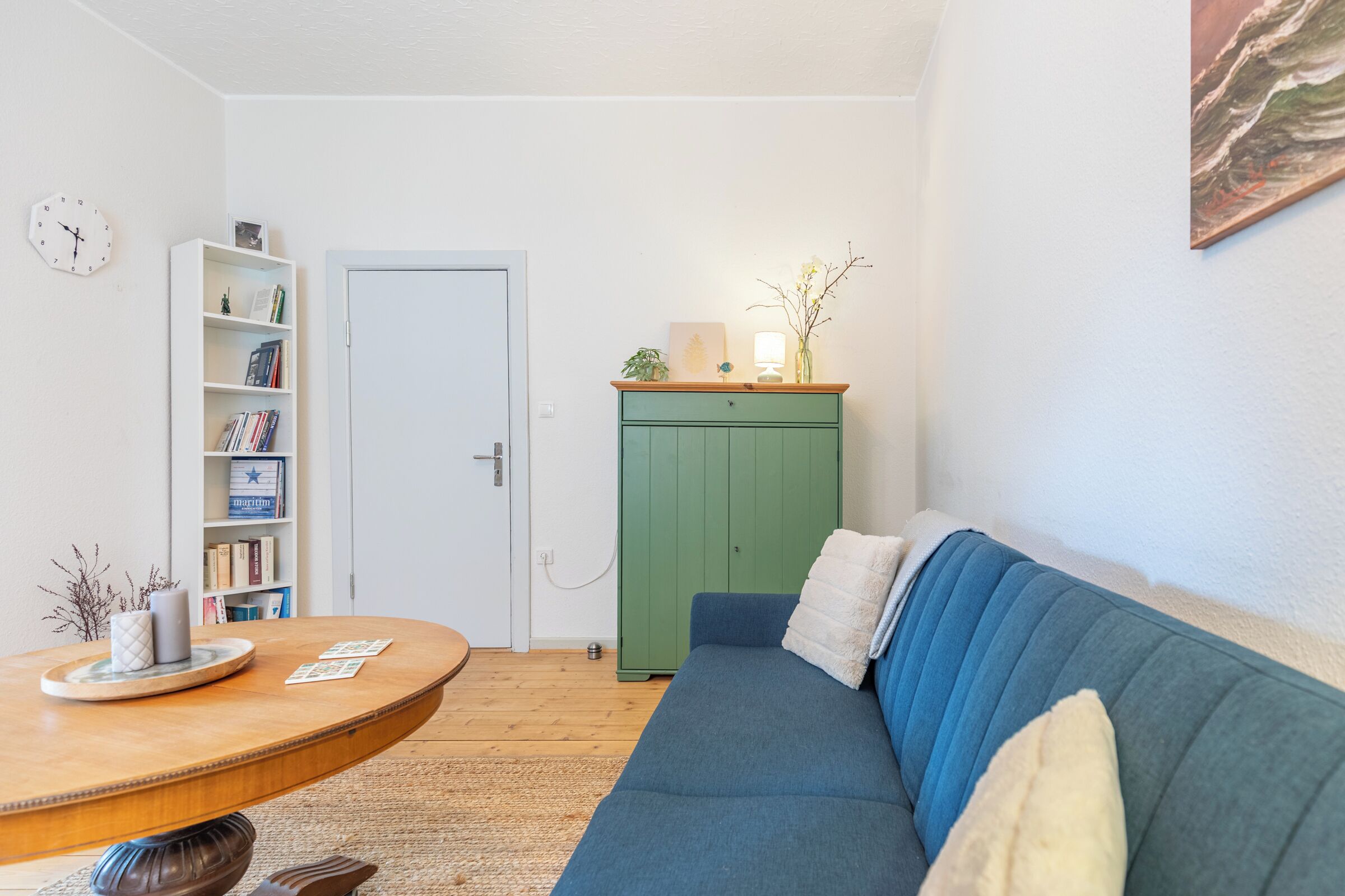 A room with a round wooden table, a blue sofa, a green cabinet, a white door, and a bookshelf.