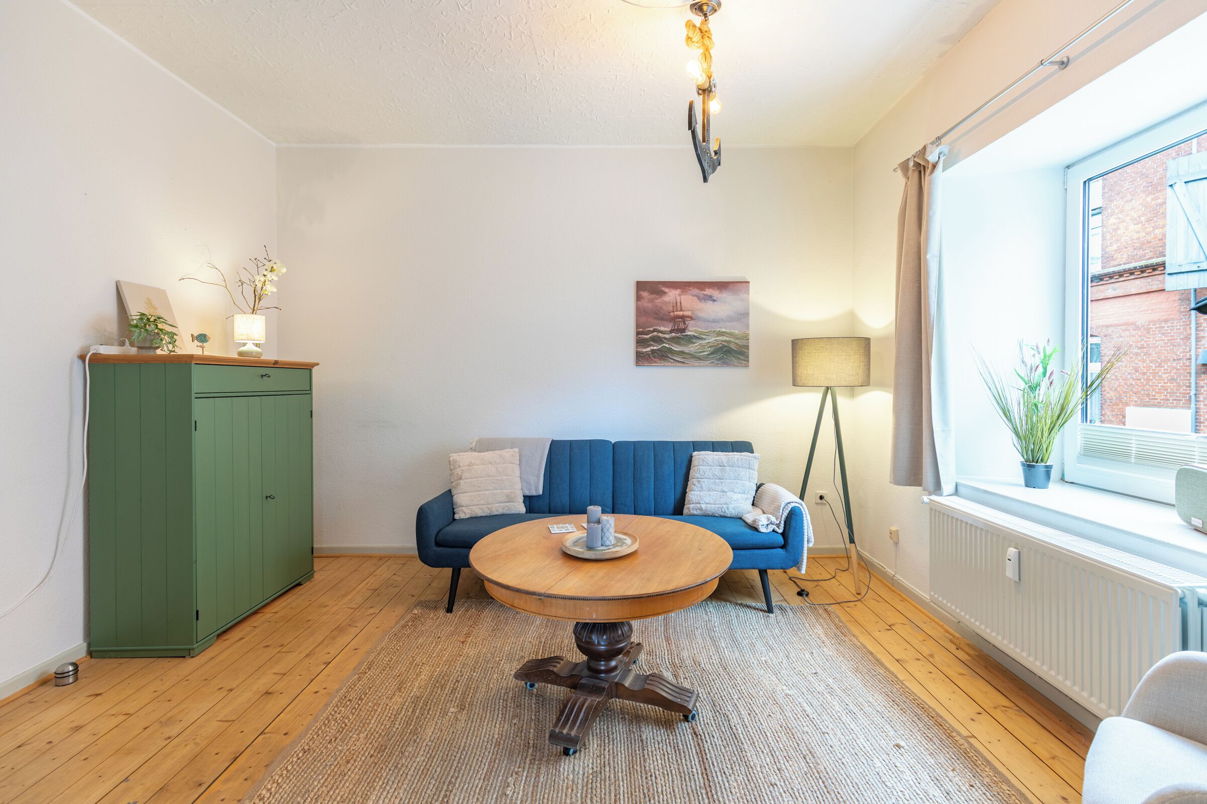 A room with a round wooden table, a blue sofa, a green cabinet, and a window with a view of a brick building.