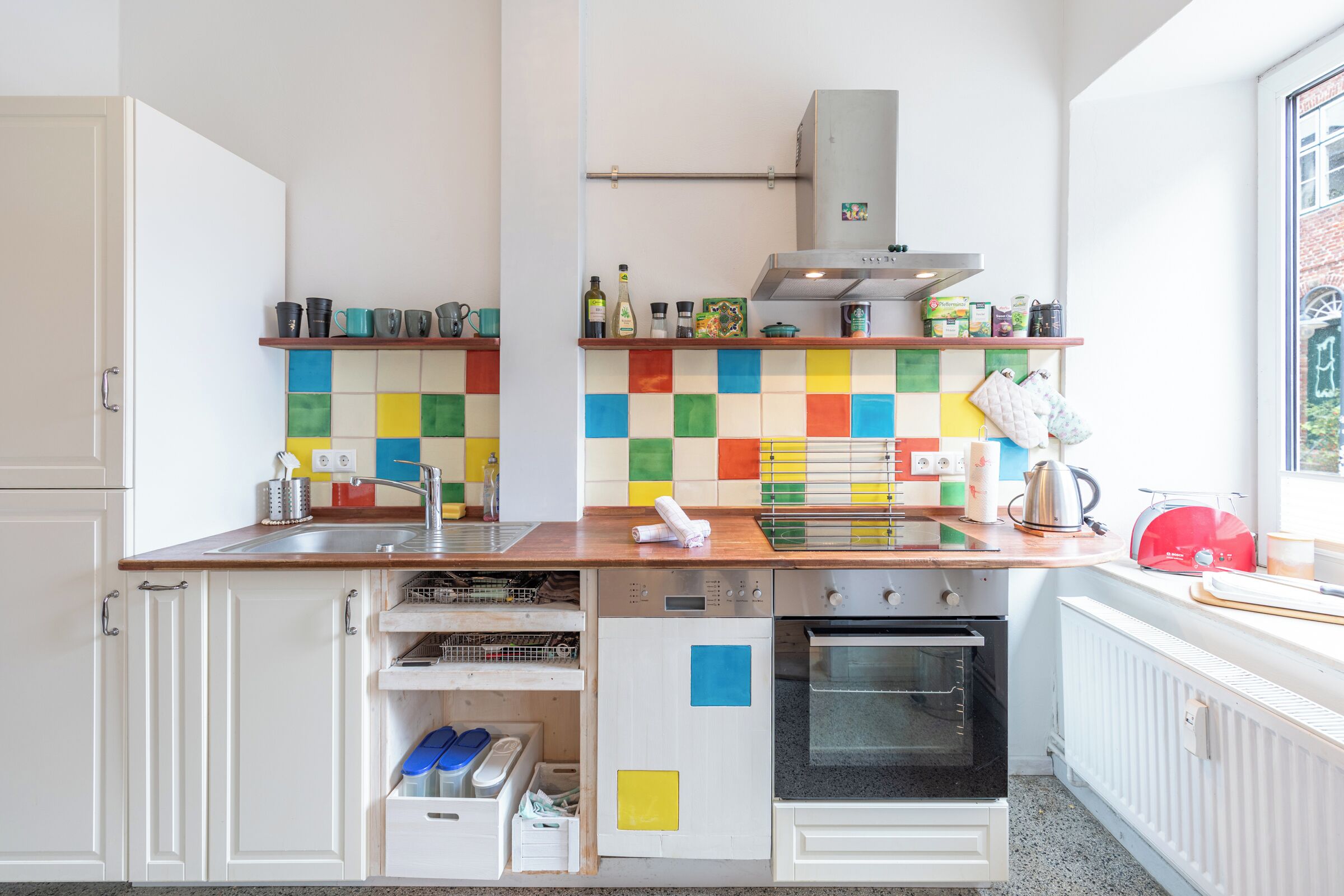 Una cocina con armarios blancos, encimera de madera, salpicadero colorido y un tostador rojo.
