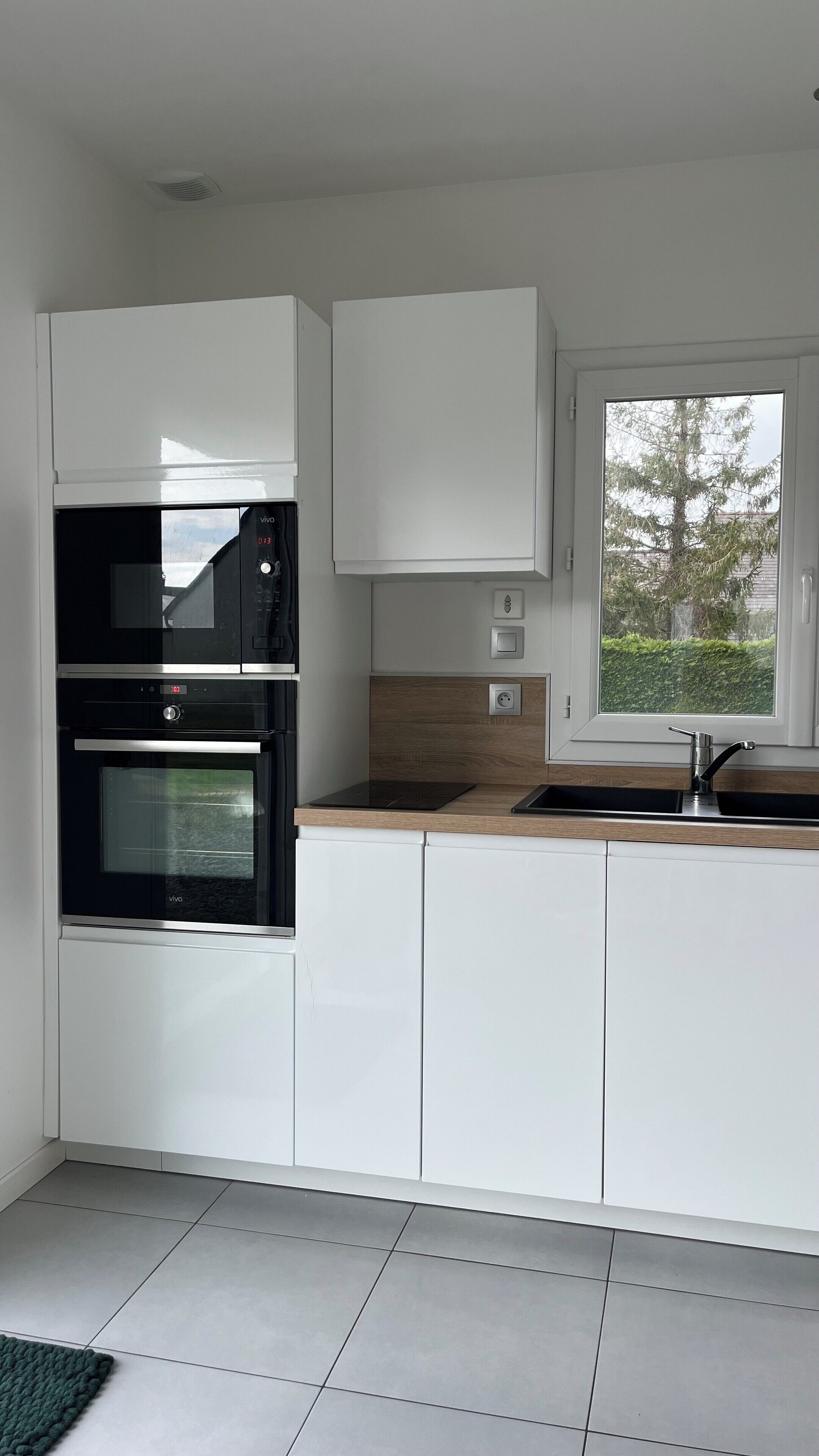 Une cuisine moderne avec des meubles blancs, un four encastré, un four à micro-ondes et une fenêtre donnant sur des arbres.
