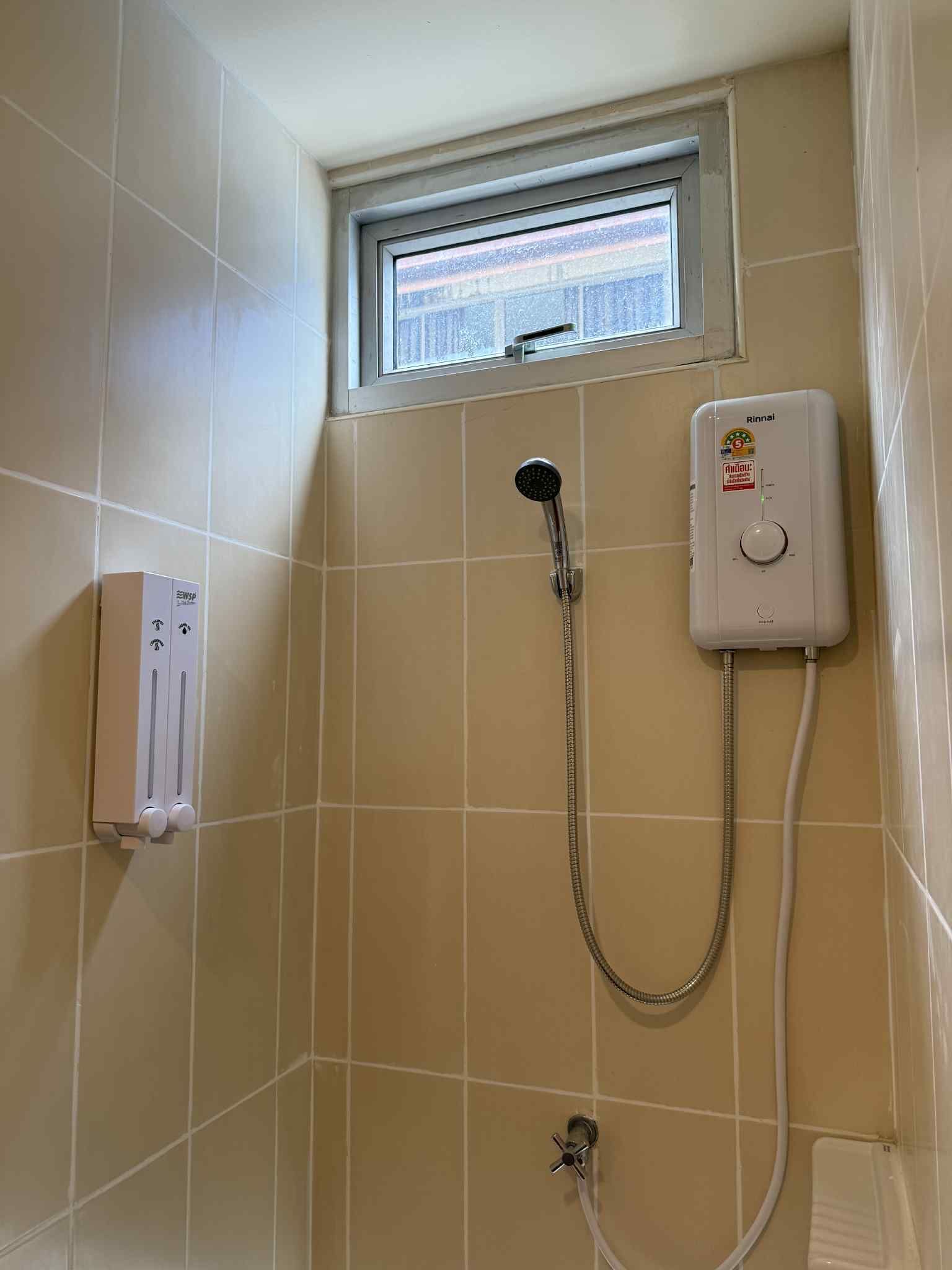 A bathroom with beige tiles, a window, a handheld showerhead, a wall-mounted water heater, and a towel dispenser.
