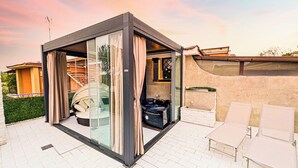 Un espace de vie extérieur moderne, doté d’une structure en verre, d’un jacuzzi et de fauteuils de salon blancs.