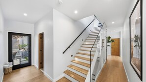 Un interior moderno con una escalera de madera, paredes blancas y un puerta de cristal que da a un espacio exterior.
