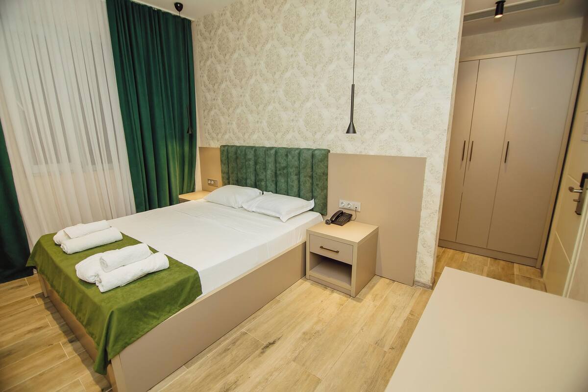A neatly made bed with a green bedspread and two white towels, a bedside table with a phone, and a wardrobe in a hotel room.