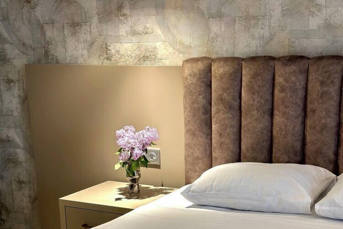 A neatly made bed with white linens, a brown headboard, and a nightstand with a vase of flowers.