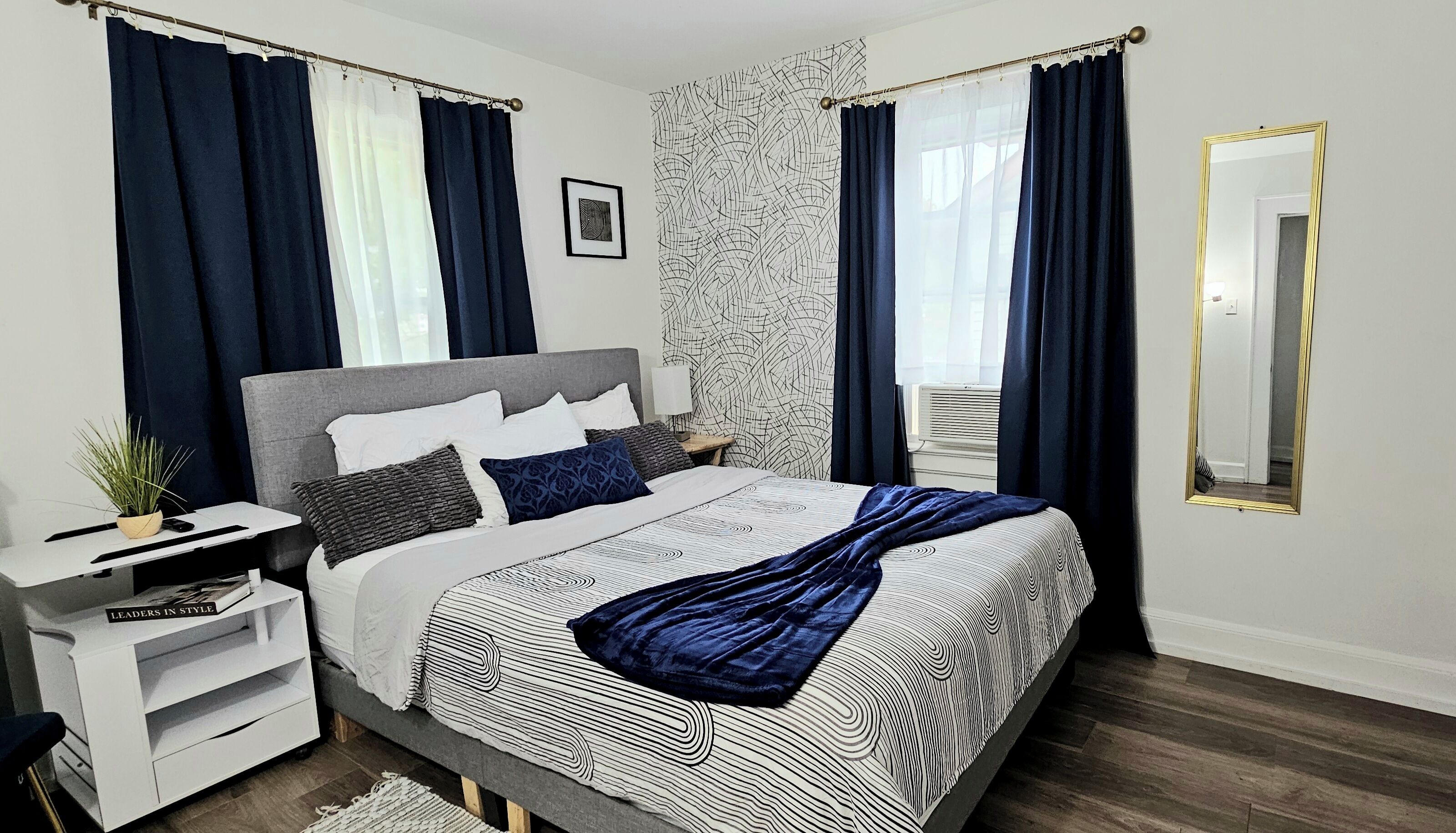 Une chambre moderne avec une tête de lit grise, un grand lit avec une literie à motifs, une table de chevet avec une lampe et un grand miroir.