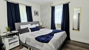 Ein modernes Schlafzimmer mit grauem Kopfteil, einem großen Bett mit gemusterten Betttextilien, einem Nachttisch mit Lampe und einem großen Spiegel.