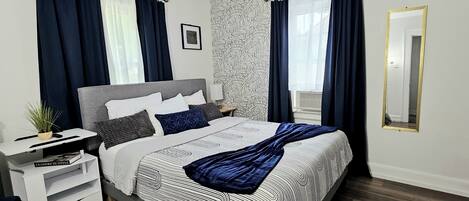 Une chambre moderne avec une tête de lit grise, un grand lit avec une literie à motifs, une table de chevet avec une lampe et un grand miroir.