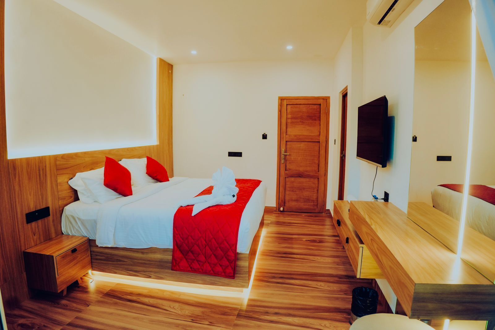 A modern hotel room with a wooden floor, a bed with white and red bedding, a wooden nightstand, a wooden bench, and a TV mounted on the wall.