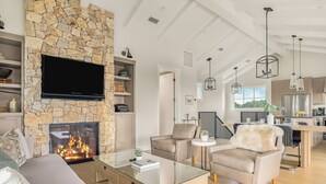 A modern living room with a stone fireplace, comfortable seating, a glass coffee table, and a kitchen in the background. - Vineyard Views w/ Pool & Game Room Near Trails | Beginnings Ranch & Vineyard (Paso Robles)