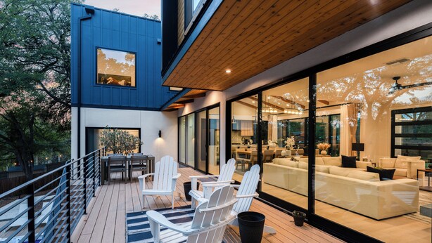 Eine moderne Außenterrasse mit weißen Adirondack-Stühlen, Holzboden und einer Glaswand, die zu einem Wohnbereich mit Sofa und Esstisch führt.