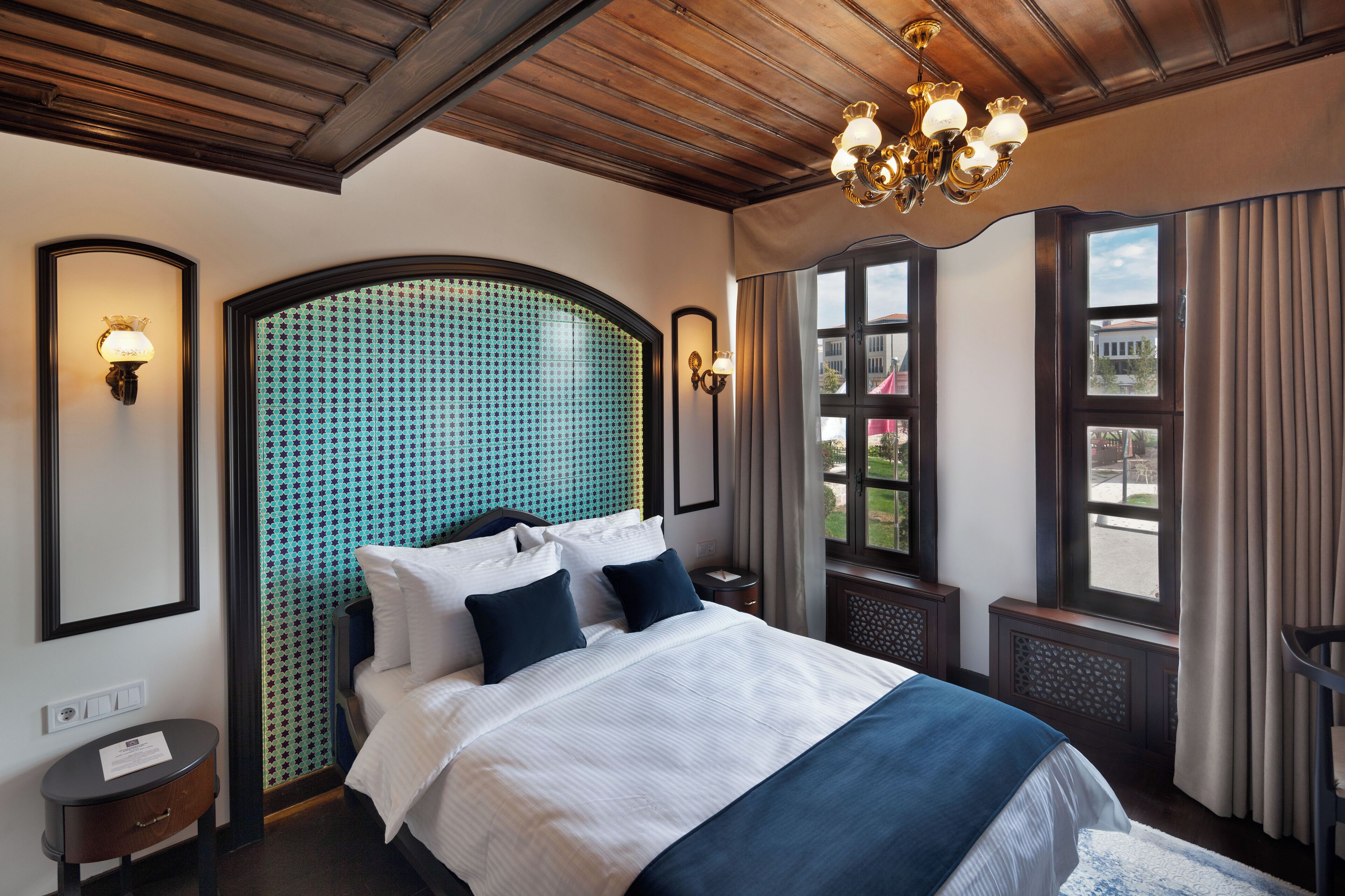 A hotel room with a large bed, a wooden ceiling, a chandelier, a window with curtains, and a wall-mounted light fixture.