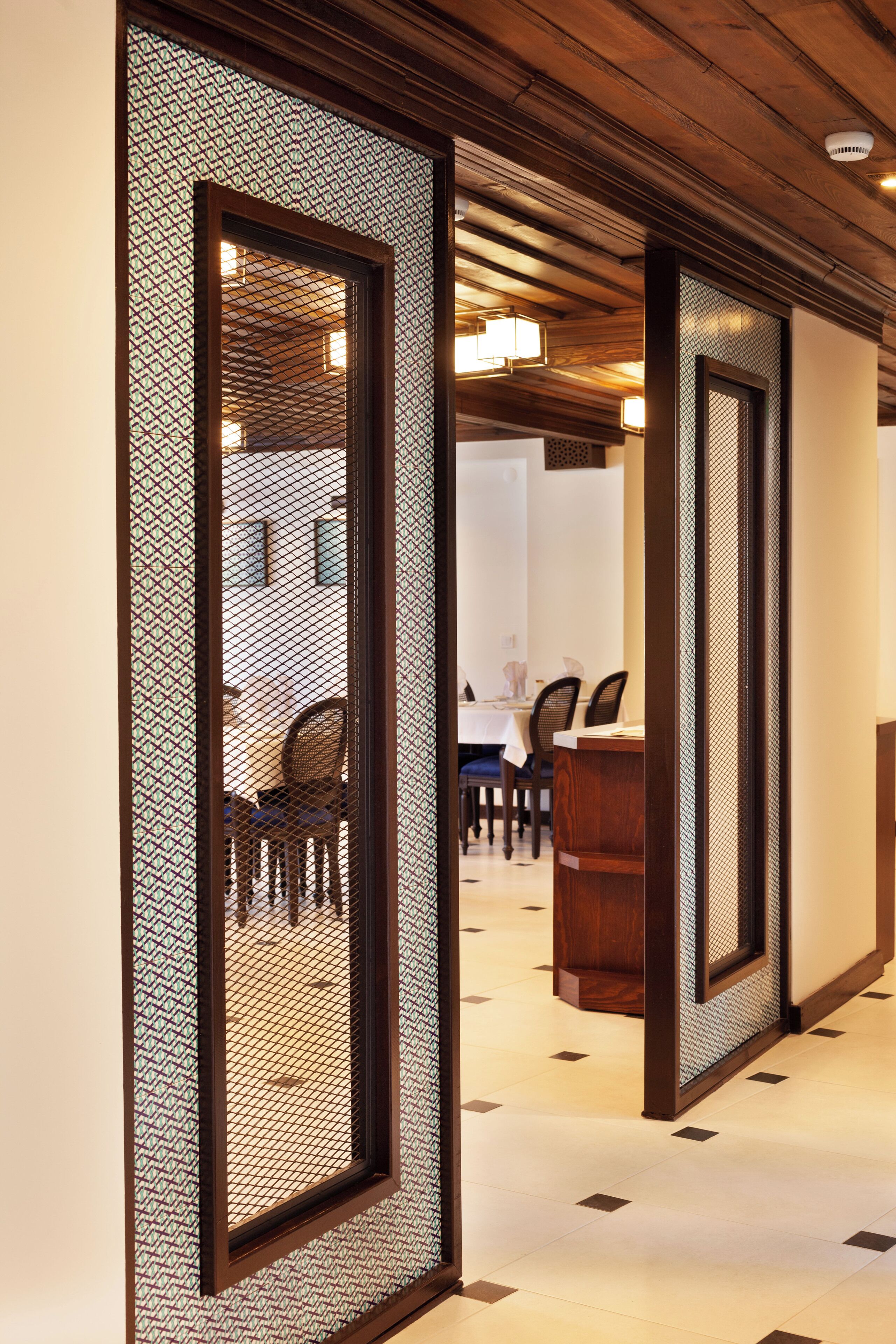 A hallway with wooden paneling and a patterned mesh design on the walls. There is a dining area visible in the background with a table and chairs.