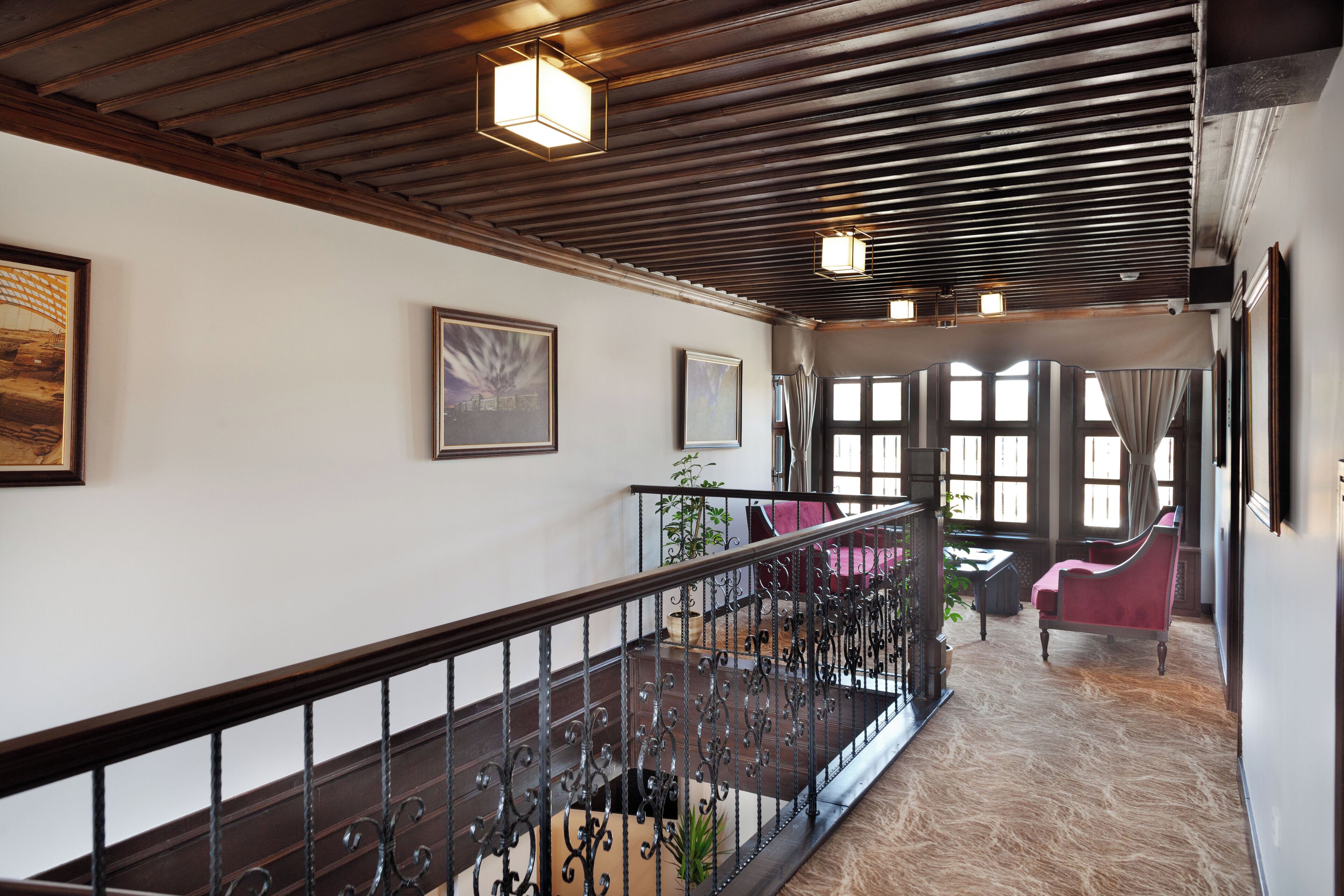 A hotel corridor with a wooden ceiling, a staircase with wrought iron railings, and a seating area with chairs and a table.
