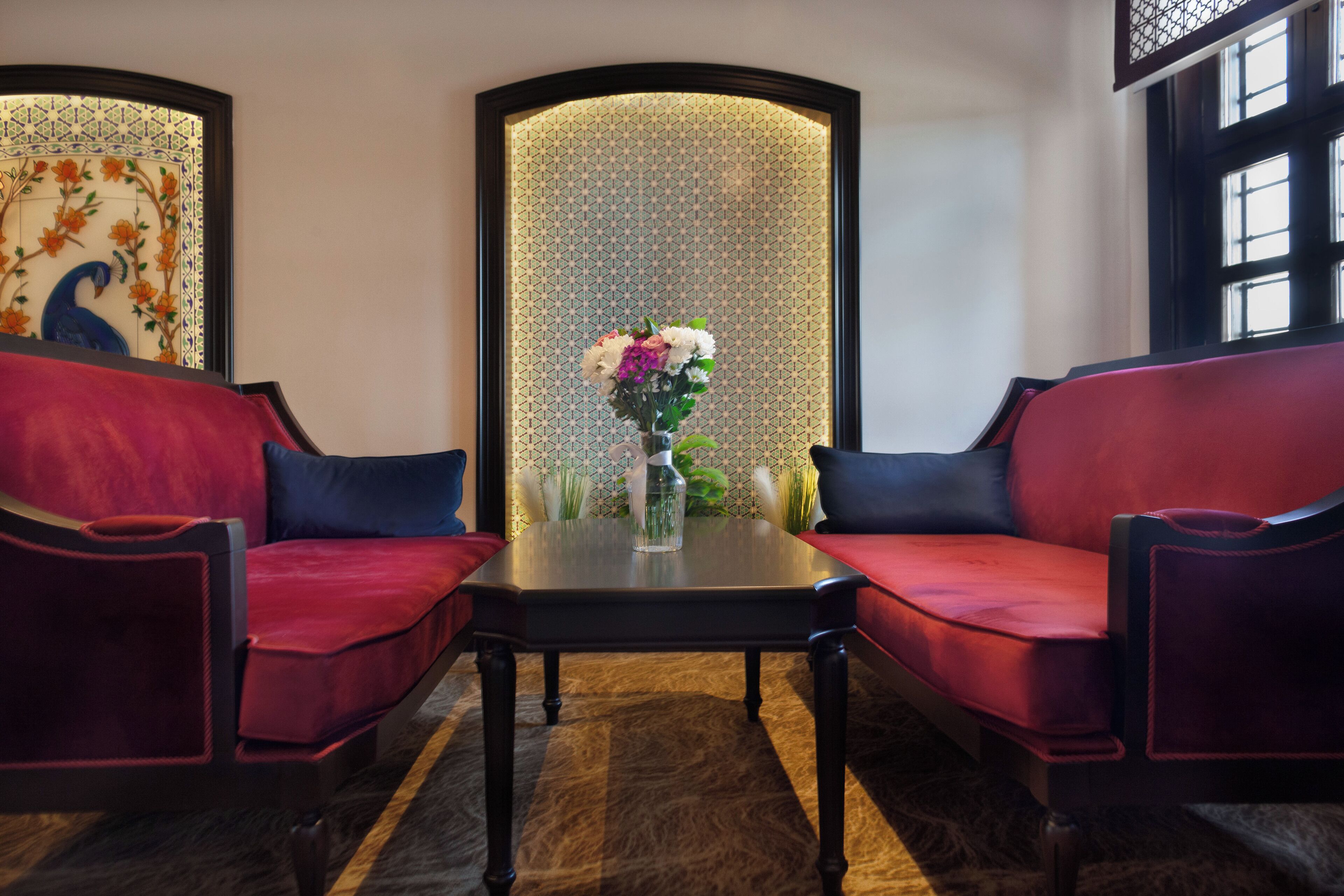 A room with two red velvet sofas facing each other, a small table with a vase of flowers, and a decorative wall with a peacock design.