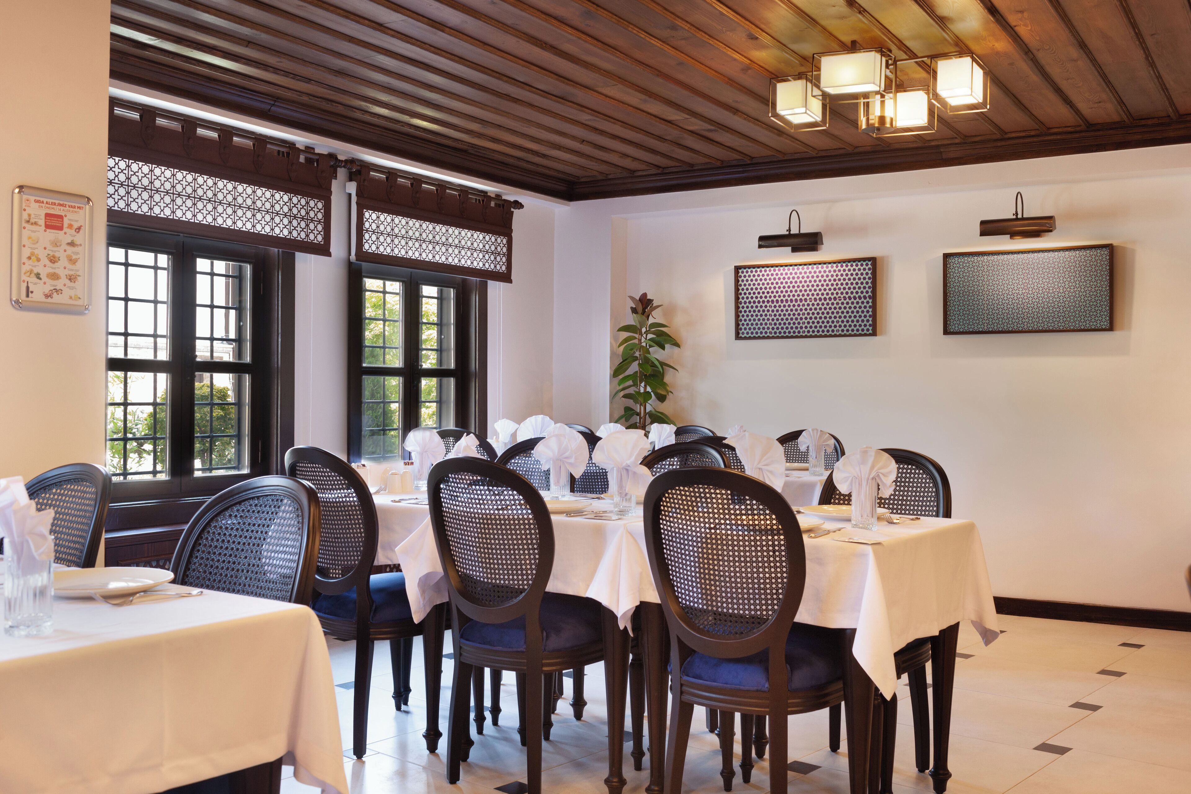 A dining area with tables set for multiple guests, featuring wooden chairs and a decorative ceiling.