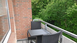 Ein Balkon mit Tisch und zwei Stühlen, eine Ziegelwand und Blick auf grüne Bäume.
