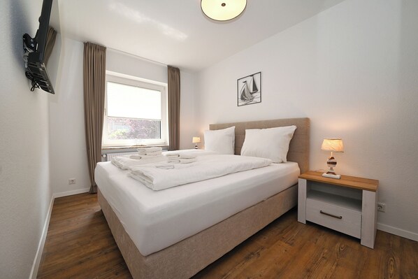 A neatly made bed with white linens, a bedside table with a lamp, and a window with curtains.
