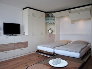 A compact bedroom with a bed, a TV, a wardrobe, and a shelf with decorative items. - House Dünenrose Apartment 6 (Norderney)