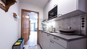Une petite cuisine équipée d’un four à micro-ondes, d’un évier et de placards. Une porte donne sur un couloir d’où l’on aperçoit une pièce.