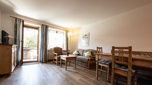 Un salon chaleureux avec un plancher en bois, un canapé, une table à manger et des chaises. On y trouve une fenêtre avec des rideaux, un radiateur et une lampe.