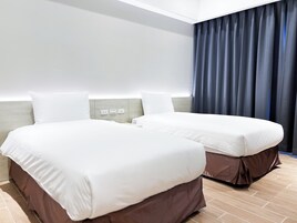 Habitación de hotel con dos camas, cabecero de madera, mesita de noche y un ventanal grande con cortinas oscuras.