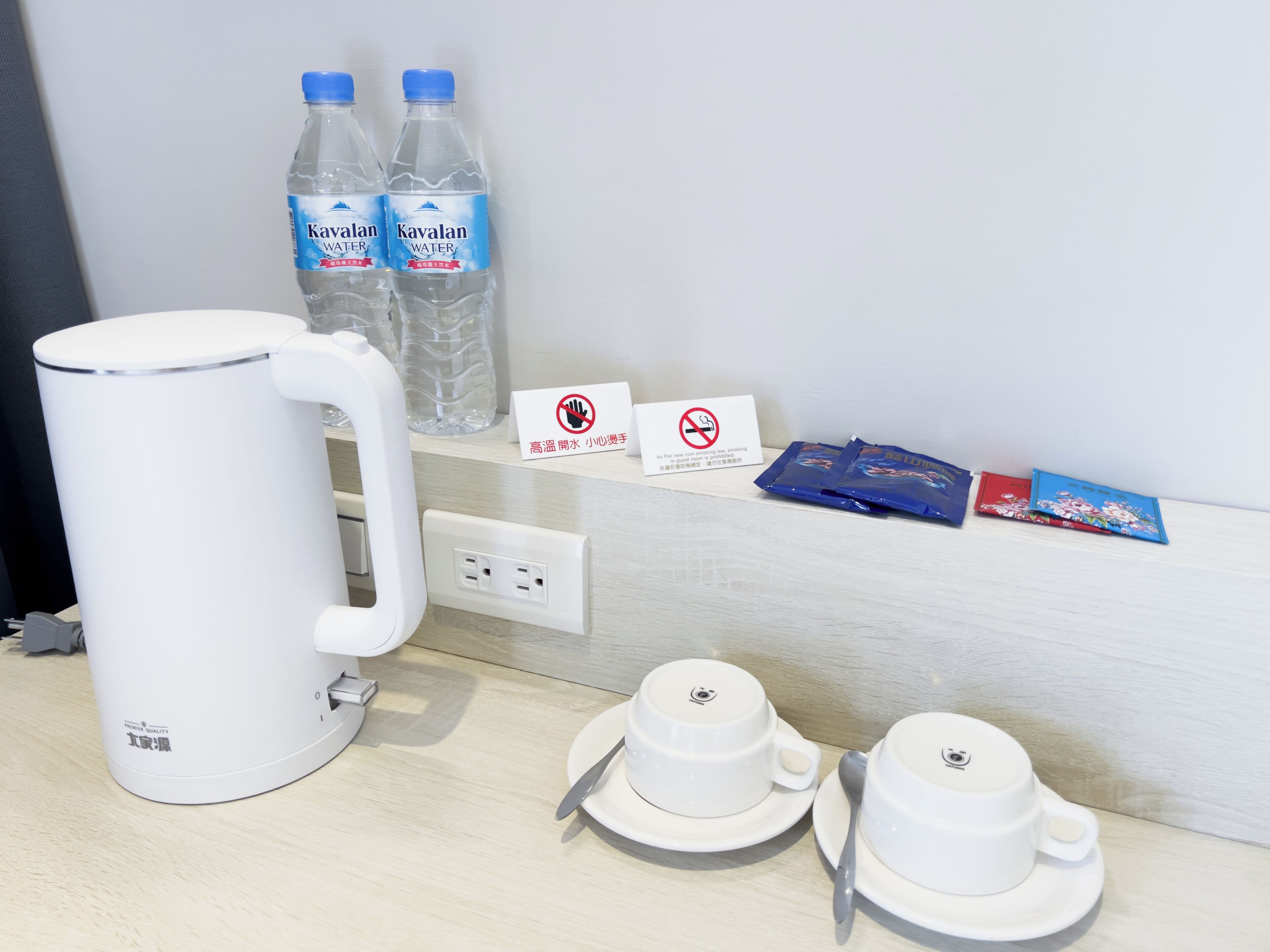 A hotel room with a white kettle, two bottles of water, a no-smoking sign, and a tray with cups and a spoon.
