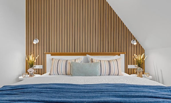 Une chambre avec un plafond en pente, une tête de lit en bois, un lit agrémenté de plusieurs coussins, des tables de chevet avec des lampes et des plantes décoratives.