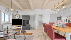 A modern kitchen with a built-in oven, a dining area with a wooden table and red chairs, and a living area with a TV and a clock. - Vacation home Marga Sophie- dreamlike idyll under thatch in Dagebüll (Dagebüll)