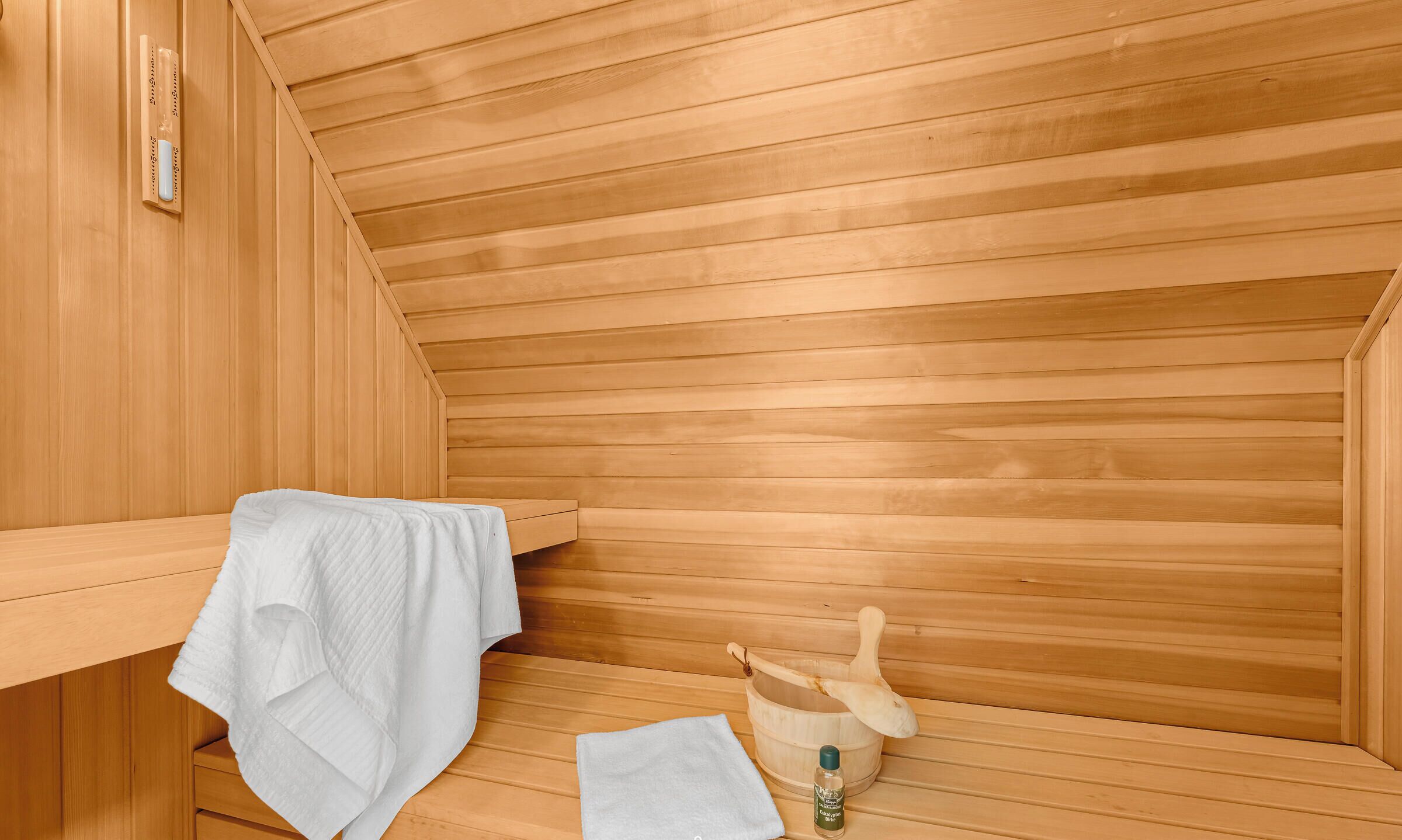 A sauna room with wooden walls, a bench, a towel, a wooden bucket, and a bottle of oil.