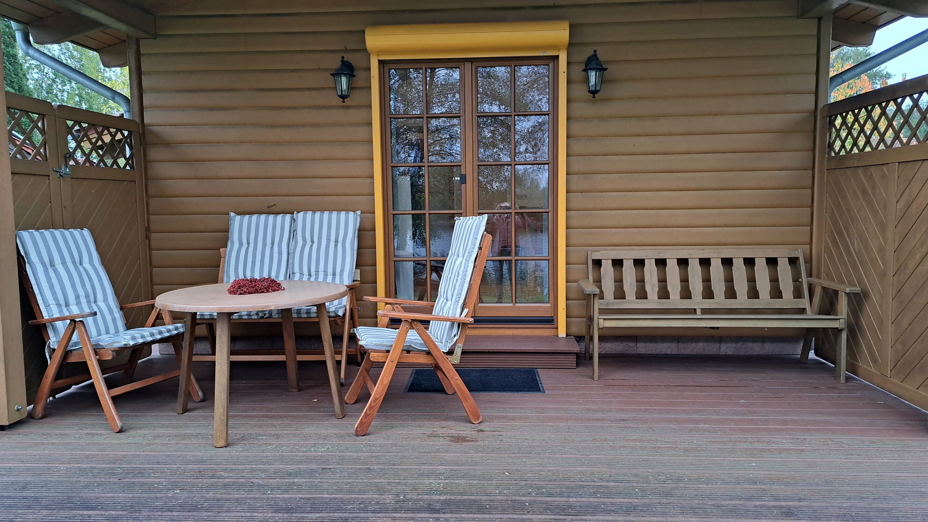 Una terraza de madera con una mesa redonda y dos sillas a rayas, un banco de madera y un ventanal con una puerta amarilla.