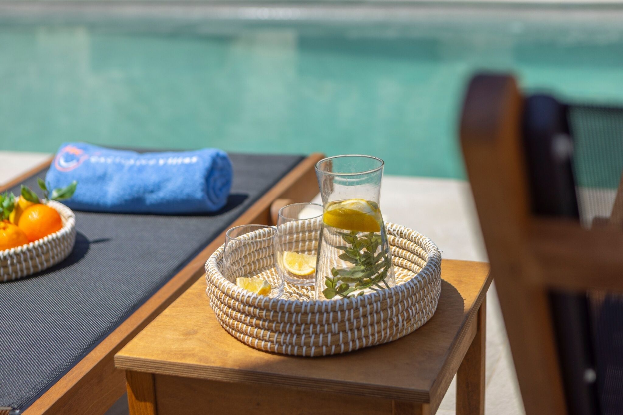 A wicker tray with glasses and lemons on a wooden table by a poolside.