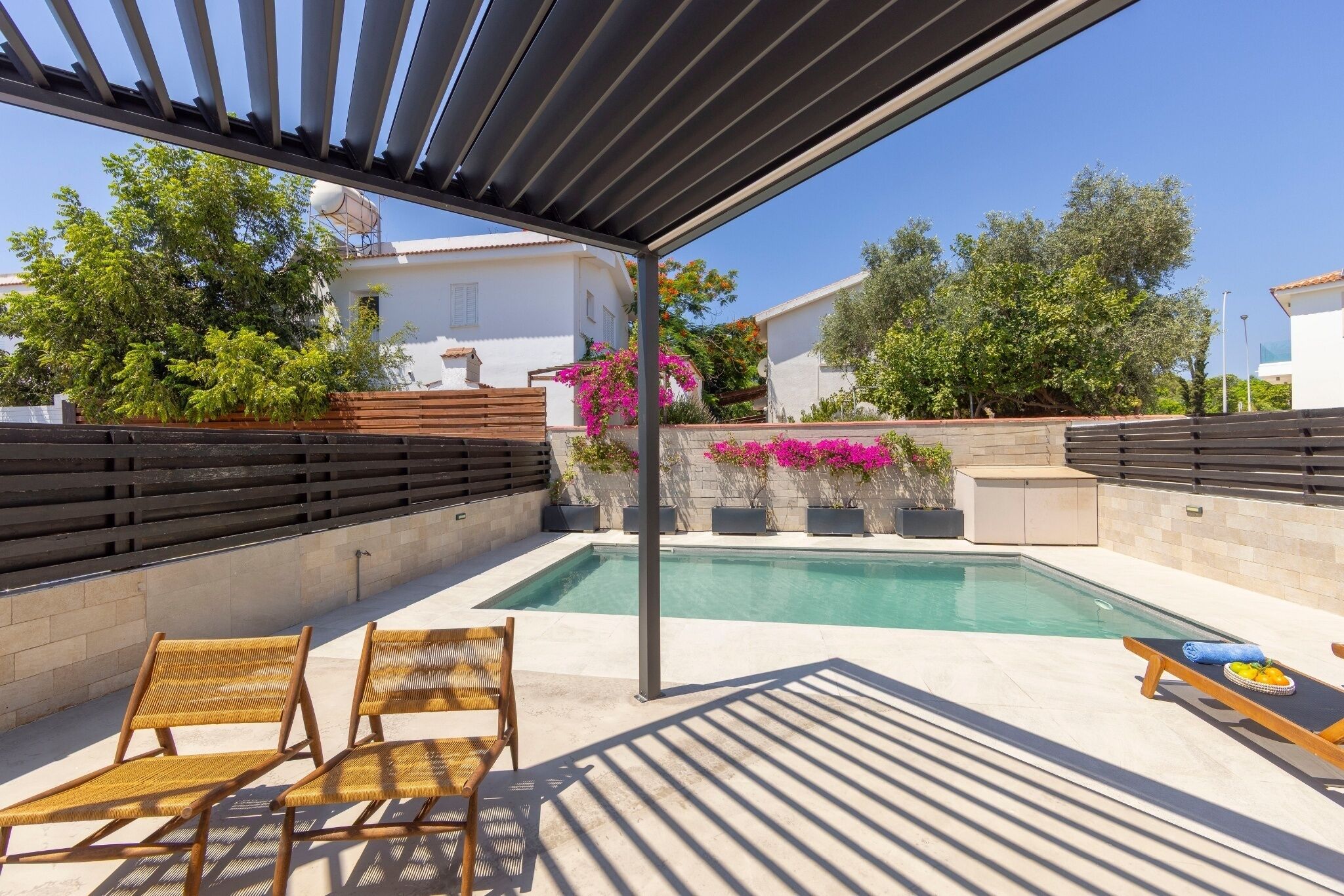 Un espacio al aire libre cubierto con piscina, sillas de madera y una mesa con frutero.