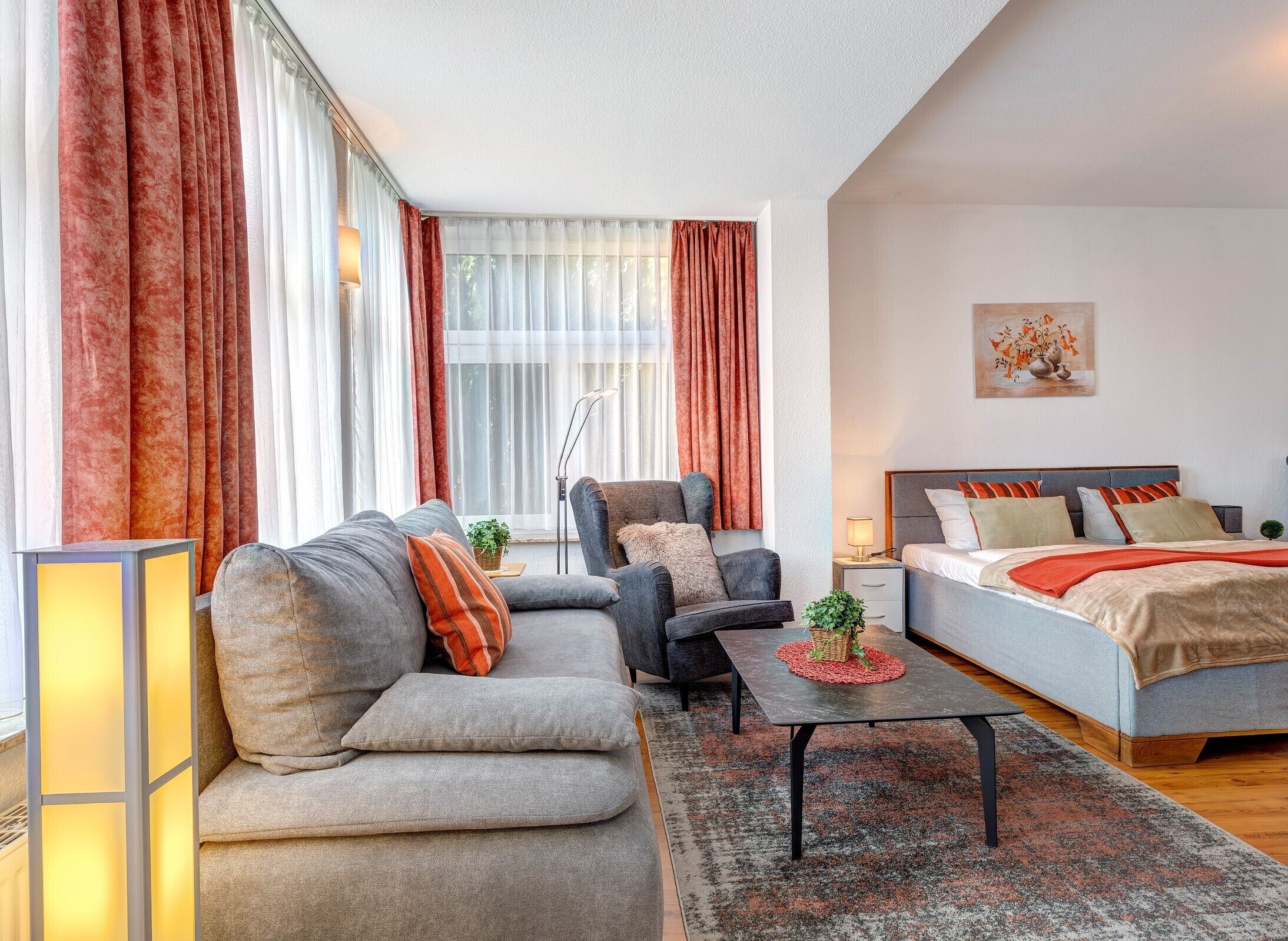 A modern living room with a sofa, armchair, and coffee table, a bed with a headboard, and large windows with curtains.