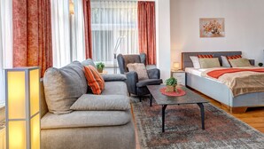 A modern living room with a sofa, armchair, and coffee table, a bed with a headboard, and large windows with curtains.